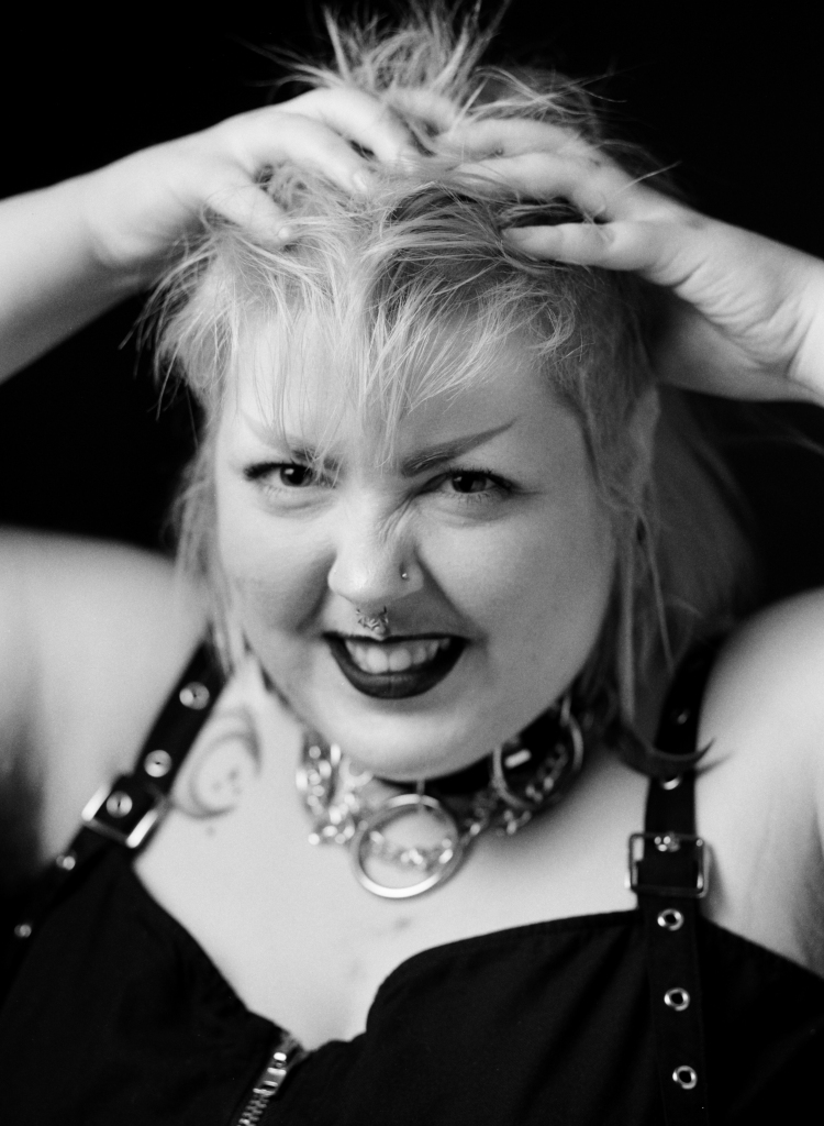 A black and white portrait of a non-binary person with a spiky mullet, piercings, a collar and dark lipstick. They are snarling at the camera in playfully with their hands in their hair. Shot on Acros 100 by Georgia Lilley in studio in Launceston, Tasmania.